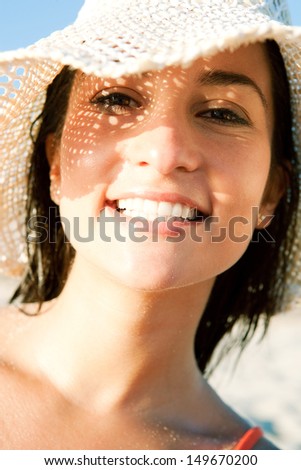 Close up portrait of an attractive smiling woman on vacation on a beach, wearing a straw hat and shading her face with it creating a sun pattern on her skin. Protecting from sun rays.