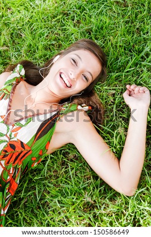 Over head portrait view of an attractive young woman laying down on textured green grass listening to music with her mp3 player and smiling joyfully during a summer day outdoors.