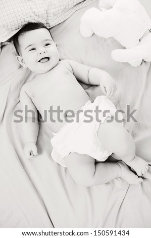 Over head black and white view of a beautiful baby girl laying down on a bed at home with her soft toy friend, joyful and smiling, and wearing a nappy.