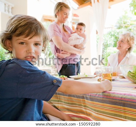 Family gathering around a table with food preparing to have lunch in the home garden, with the boy child turning to the camera during a sunny day outdoors.