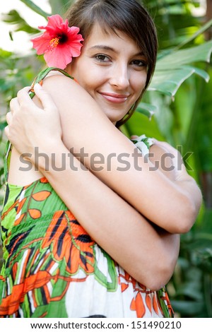 Beauty portrait of a beautiful young woman hugging herself in a lush forest with tropical leaves in an exotic travel destination, smiling with a red flower in her hair, outdoors.