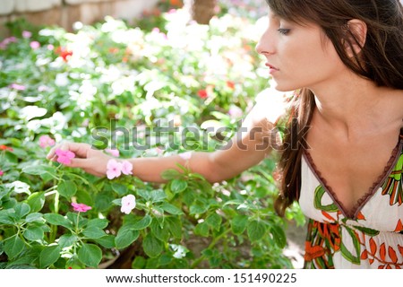 Side view of a young attractive woman in a green park stretching her arm to pick a flower from a bush full of them, being selective and focused, outdoors.