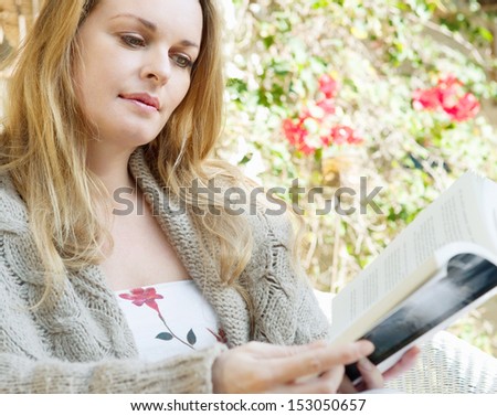 Portrait of an attractive woman reading a book while relaxing in a home garden on vacation, sitting down on a wicker chair during a sunny day, exterior.