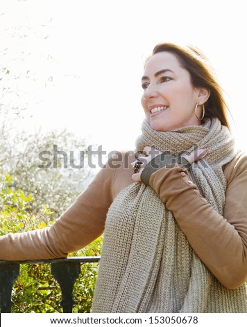 Portrait of a middle aged attractive woman smiling and wearing a thick woolly knitted scarf while leaning on a banister in her country home during a sunny autumn day.