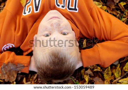 Over head close up portrait of a blond child boy laying down on a bed of autumn dry leaves during the fall, looking up at camera and relaxing during a day out in nature.