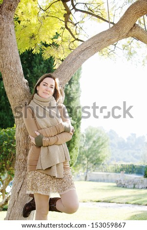 Elegant and sophisticated middle age attractive woman leaning on a tree trunk in her home garden, smiling and enjoying a sunny autumn day during the fall season, outdoors.