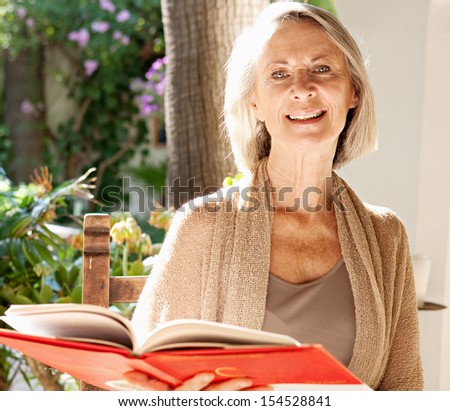 Active mature attractive woman sitting and relaxing in a home garden holding and reading a book, smiling during a sunny day outdoors.