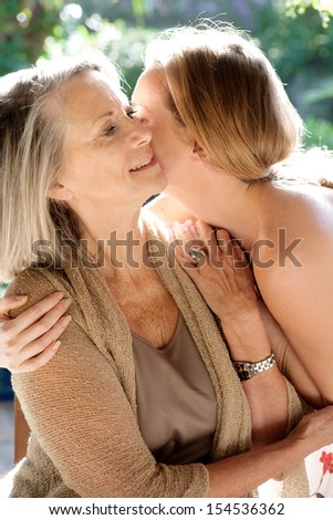 Mature mother and adult daughter spending time together in a home garden whispering and kissing during a bright and golden summer sunny day.