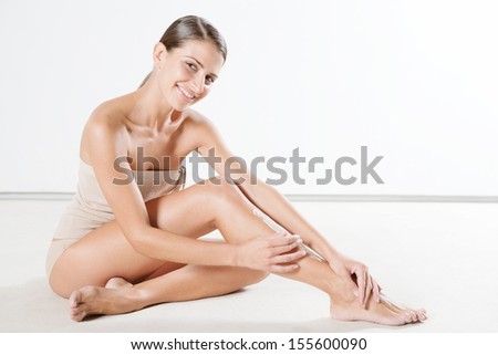 Attractive young hispanic woman applying hydrating lotion cream on her legs tanned skin while sitting down against a white background smiling, indoors.