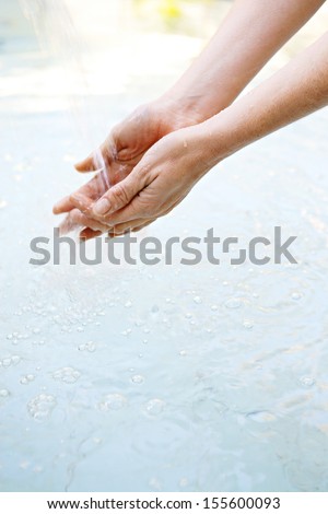 Close up view of a woman hands held together under the falling cleansing water of a spa fountain during a sunny day outdoors.