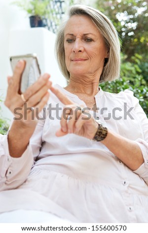 Attractive mature woman relaxing on a green garden and sitting on a white sofa holding and using a smartphone mobile device with touch screen, outdoors.
