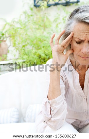 Mature woman sitting on a white sofa in a home garden touching her head with her hand and fingers while having a headache pain and feeling unwell, outdoors.