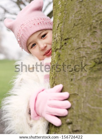 Young child girl playing and hiding behind a tree trunk and hugging it in a green park during a winter cold day, outdoors.