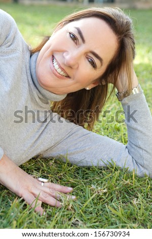Portrait of an attractive hispanic middle aged mature woman laying down on the green grass of a home garden relaxing and leaning on her hand, joyful and smiling, outdoors.
