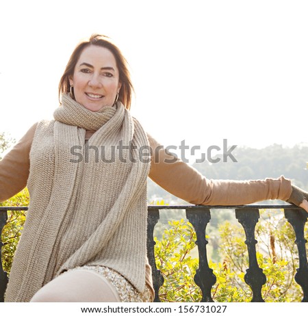Portrait of an attractive hispanic middle aged mature woman in a home garden relaxing and leaning on an iron balcony banister, looking at camera and smiling with space, outdoors.