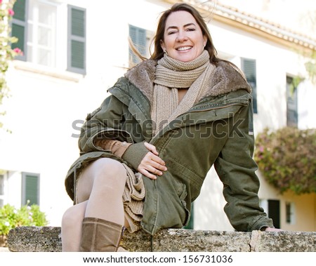 Attractive middle aged mature woman sitting at a rural home garden stone wall during a winter morning, relaxing and enjoying the sunny day wearing a big and warm coat and boots.