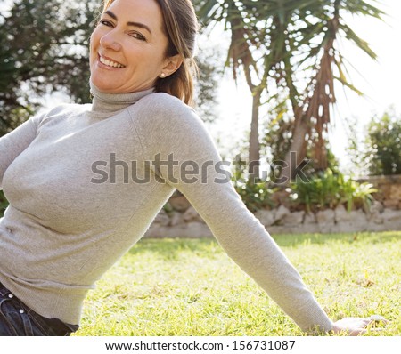Portrait of an attractive hispanic middle aged mature woman relaxing on the green grass of a home garden during a sunny fall day, joyful and smiling outdoors.