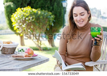 Close up portrait of an attractive middle aged mature woman relaxing and reading a book in a country home garden during a sunny autumn morning, near a breakfast table, smiling.