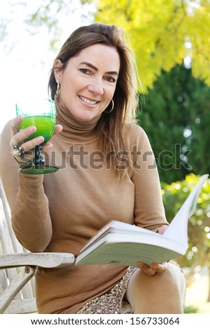 Close up portrait of an attractive middle aged mature woman smiling and relaxing reading a book in a country home garden during a sunny autumn morning, outdoors.