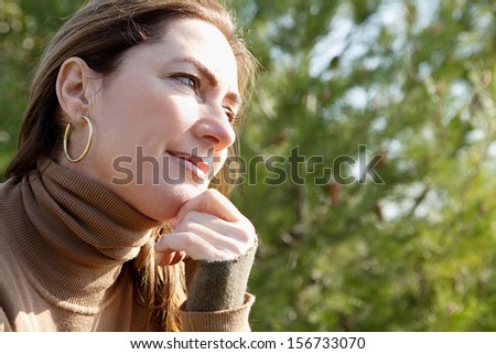 Close up portrait of an attractive middle aged mature woman visiting the countryside and leaning on her hand being thoughtful during a sunny winter morning outdoors.