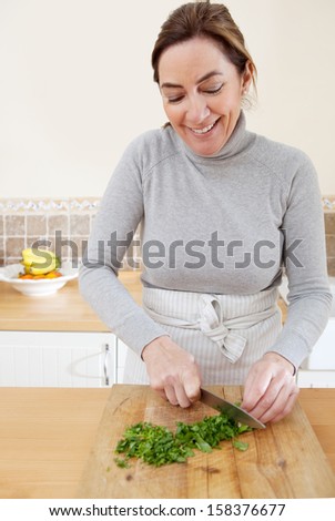 Attractive mature woman chopping parsley herbs and cooking vegetables in the kitchen at home, with healthy organic produce and using a knife and chopping board, smiling.