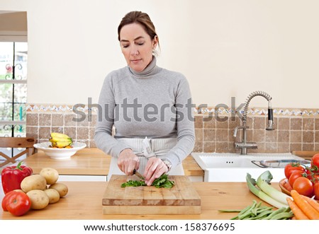 Attractive mature woman chopping herbs and cooking vegetables in the kitchen at home, with healthy organic produce and using a knife and chopping board.