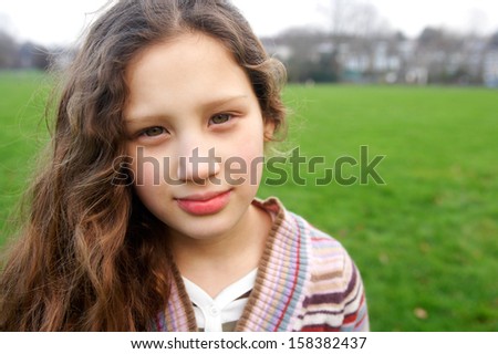 Close up portrait of a beautiful young girl with a gentle smiling expression, wearing a stripy knitted jumper while in a green grass park field during a winter autumn day outdoors.