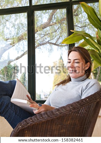 Attractive mature woman with her feet up on a table, sitting in a home conservatory with large glass windows and a green garden, reading a book and relaxing indoors, smiling.