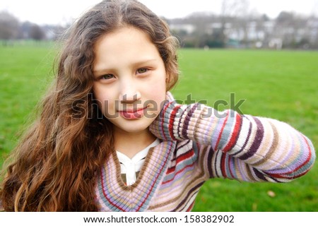 Close up portrait of a beautiful young girl with a gentle smiling expression, wearing a red knitted stripy jumper while in a green grass field park during winter autumn day outdoors.