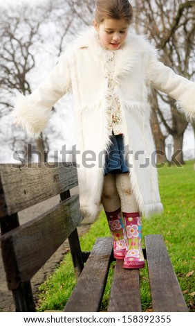 Young child girl being playful and walking slowly on a wooden bench in a green park, keeping her balance while wearing wellington boots and a coat during a cold winter day, outdoors.