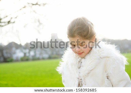 Beautiful young girl child wearing a coat during a cold winter day in a park with green grass and a sunny sky, looking down and being thoughtful, outdoors.