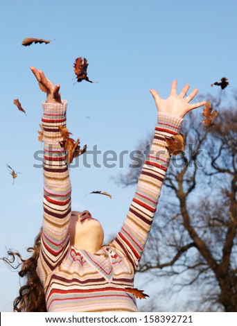 Young girl throwing dry autumn leaves up into the blue sky with her arms and and with floating leaves falling against a bright blue sky and leafless fall trees, outdoors.