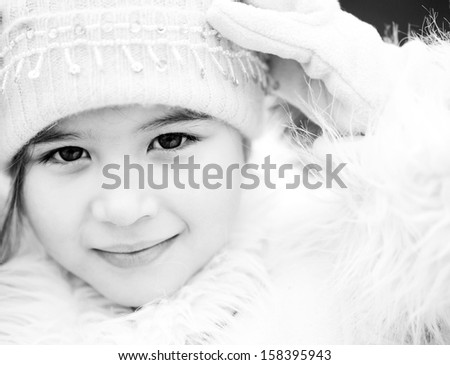 Black and white close up beauty portrait of a young child girl wearing a winter coat, woolly hat and gloves, smiling to the camera during a cold winter day, outdoors.
