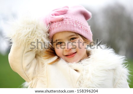 Young beautiful girl with her hand holding her woolly hat while visiting a park during a winter day and wearing a warm coat, smiling outdoors.