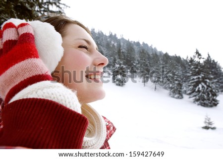 Close up side beauty portrait of a smiling young woman breathing fresh air in the snow mountains, feeling healthy and wearing ear warmers during a cold winter day outdoors.