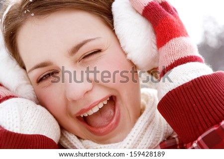 Close up portrait of a joyful and attractive woman wrapped up during winter and holding her ear warmers while laughing and winking at the camera on vacation in the snow, outdoors.
