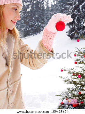 Side close up portrait of an attractive and smiling blond woman in the snow mountains nature, decorating a Christmas tree with bar balls and gifts holding a ball up during a winter day.
