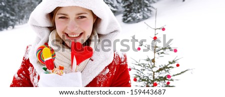 Panoramic view of an attractive young girl dressed as Santa Klaus in the snow mountains, decorating a Christmas tree with bar balls and holding a x-mas sock full of sweets, joyfully smiling.
