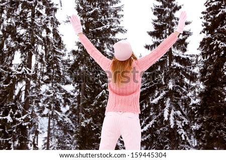 Rear view of a young woman with blond hair and a pink jumper, hat and gloves with her arms raised up in the air, enjoying the snow trees in the winter mountains, outdoors.