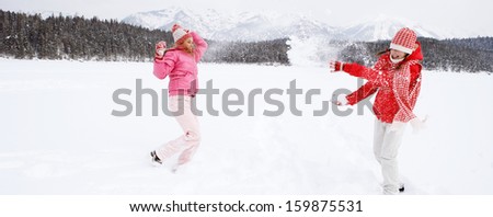 Panoramic view of two joyful and energetic friends playing games and having fun, having a snow ball fight in the snow mountains landscape during a skiing holiday on a sunny winter day.