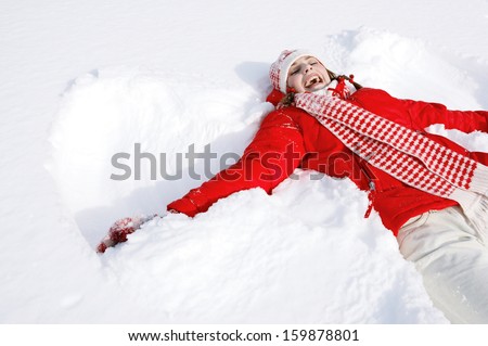 Portrait of a beautiful young woman laying down on a frozen snow lake moving her arms and legs up and down creating a snow angel figure, playing games during a sunny winter  vacation.