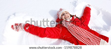 Beautiful joyful young woman laying down on a frozen snow lake moving her arms up and down creating an angel figure shape, playing games while on a skiing vacation, panoramic format.