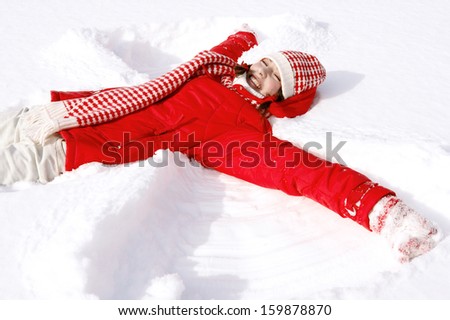 Side view of a beautiful young woman laying down on a frozen snow lake moving her arms up and down creating an angel figure shape, playing games while on vacation during a sunny winter day.