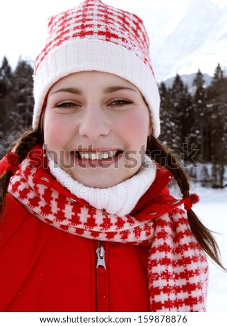 Close up portrait of a beautiful young woman on vacation in the snow mountains, smiling at the camera and wearing red winter clothing during a sunny cold day, outdoors.