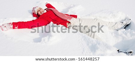 Panoramic side view of a young woman laying down on a frozen snow lake moving her arms and legs up and down creating an angel figure shape, playing games during a sunny winter  vacation.