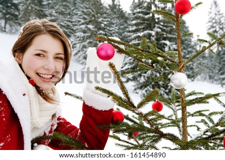 Portrait of an attractive young woman having fun and playing in the snowed mountains, decorating a small Christmas tree with bar balls, while joyfully smiling in the snow, outdoors.