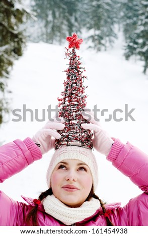Portrait of an attractive young woman having fun and playing in the snowed mountains, balancing a small Christmas tree on top of her head, joyfully smiling in the snow, outdoors.