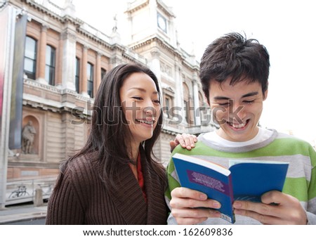 Portrait of a young and attractive Japanese tourist couple visiting the city of London on holiday together and reading a guide book in a classic street, laughing joyfully outdoors.