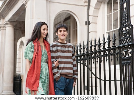 Young Japanese couple enjoying a holiday trip to the city of London together, sightseeing and walking passed a railing in a classic stone buildings street, smiling outdoors.