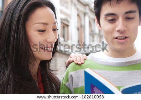 Close up portrait of a young and attractive Japanese tourist couple visiting the city of London on holiday together and reading a guide book in a classic street, laughing joyfully outdoors.